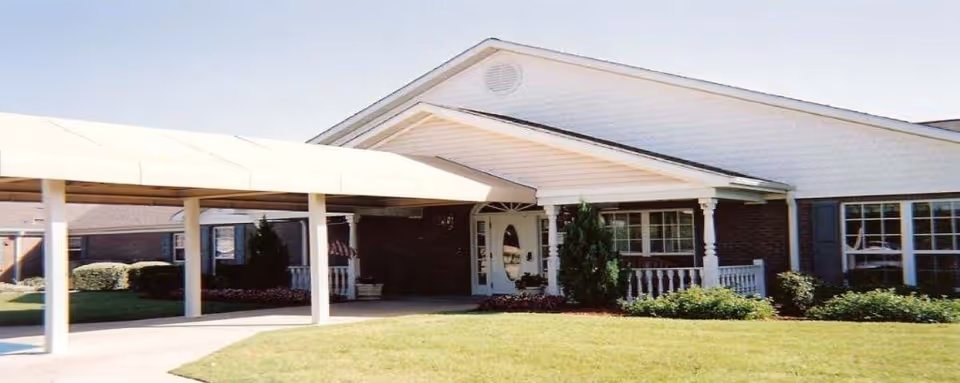 Front exterior of a single-story memory care building with a covered drive-up entrance, porch, and lawn.