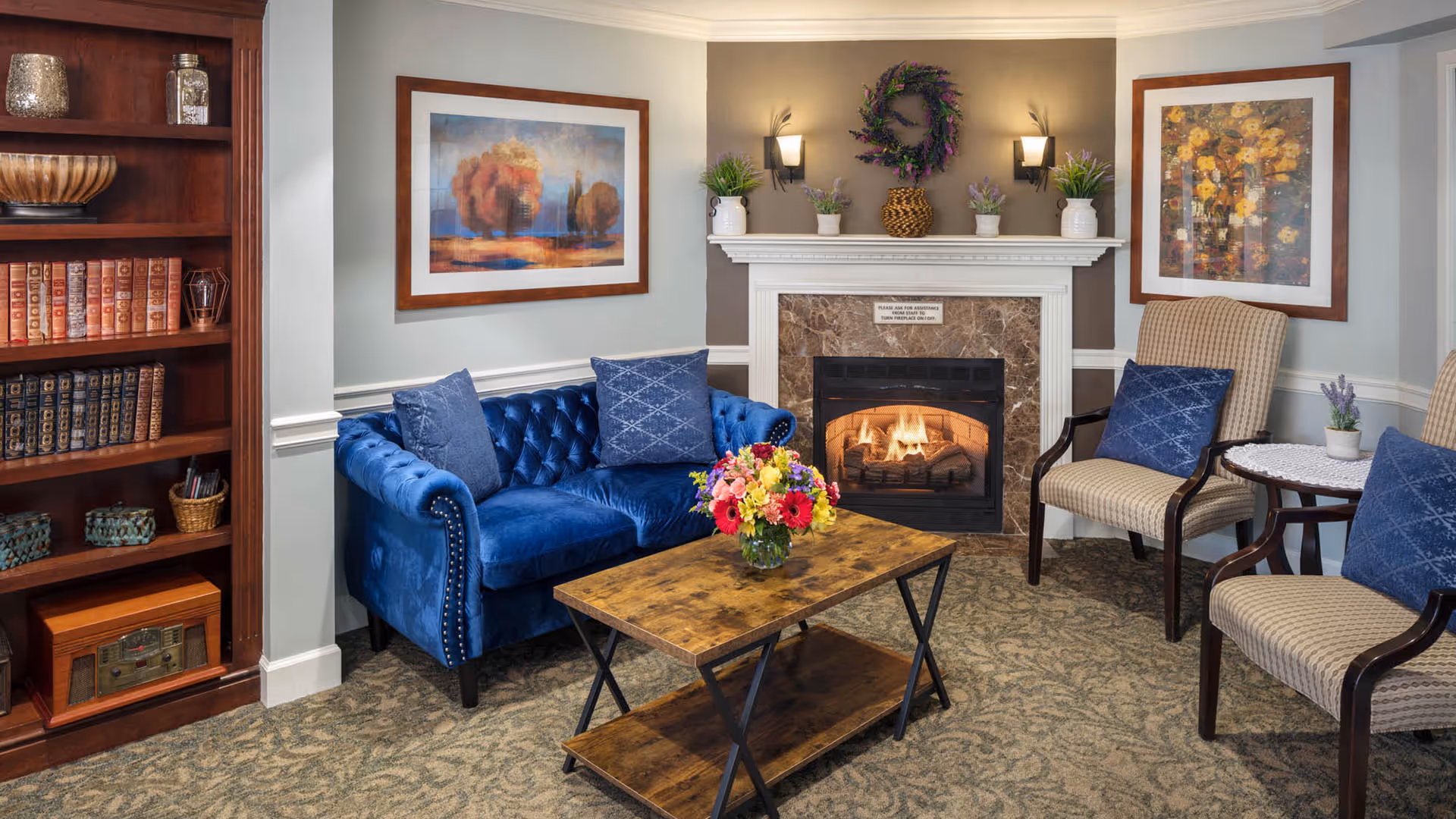 A cozy living room area with a blue velvet tufted sofa, two beige armchairs with blue cushions, a wooden coffee table with a colorful flower arrangement, a fireplace with a lit fire, and decorative plants and artwork on the walls.