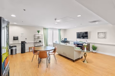 A bright and spacious common area in a senior living facility featuring a round wooden table with four brown chairs, a beige sofa facing a wall-mounted flat screen TV, a small side table with a vase of flowers, a shelving unit with decorative items, and a window with green curtains letting in natural light. The room has light wood flooring and white walls.