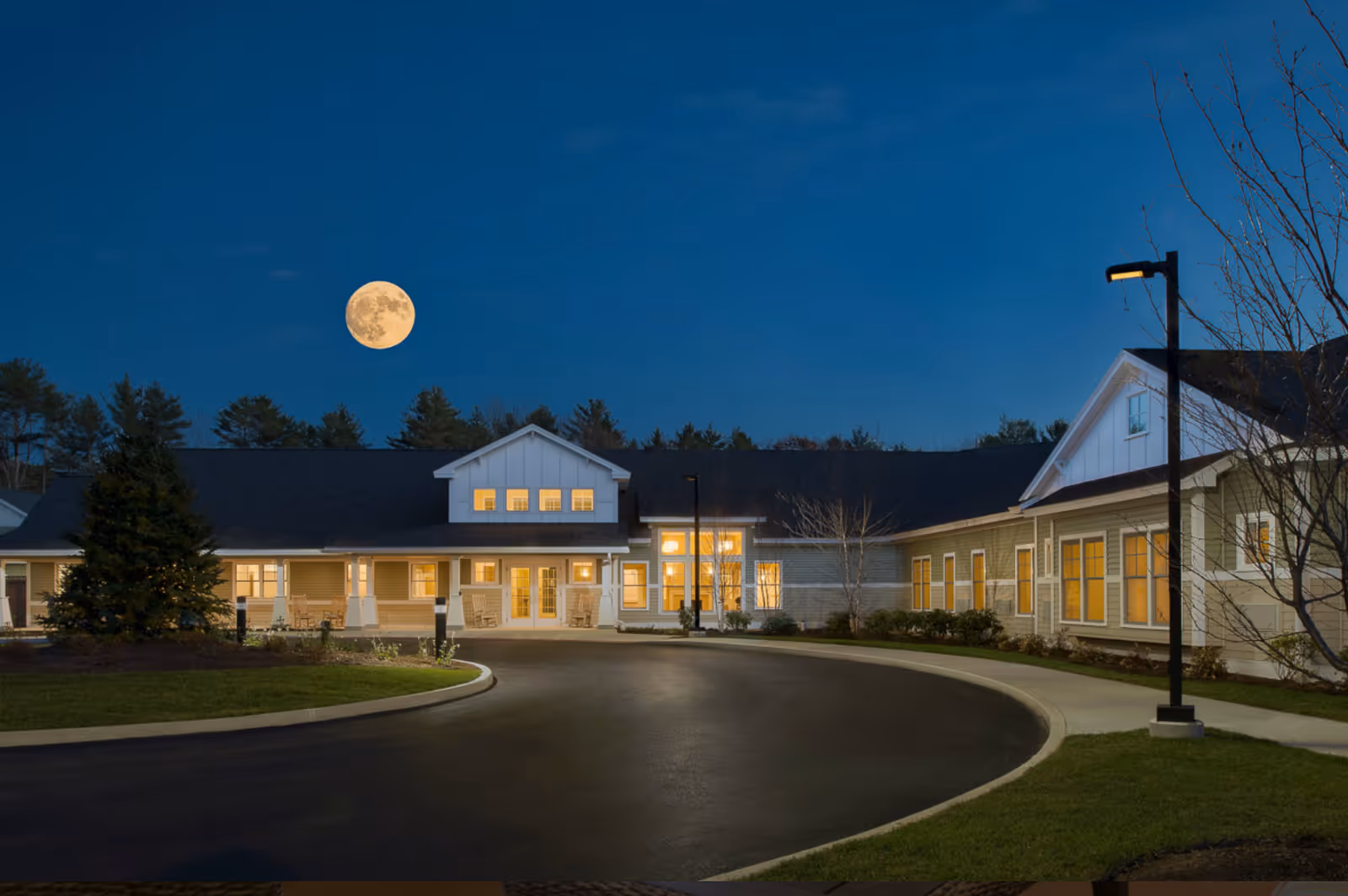 Exterior view of Avita of Brunswick senior living facility at dusk with a full moon in the sky, illuminated windows, a curved driveway, and a lit streetlamp.