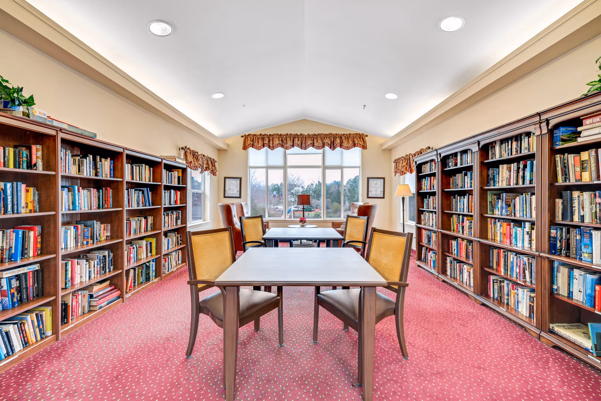 Bright communal library/reading room with bookshelves along both walls, tables and chairs, and a large window at the far end.