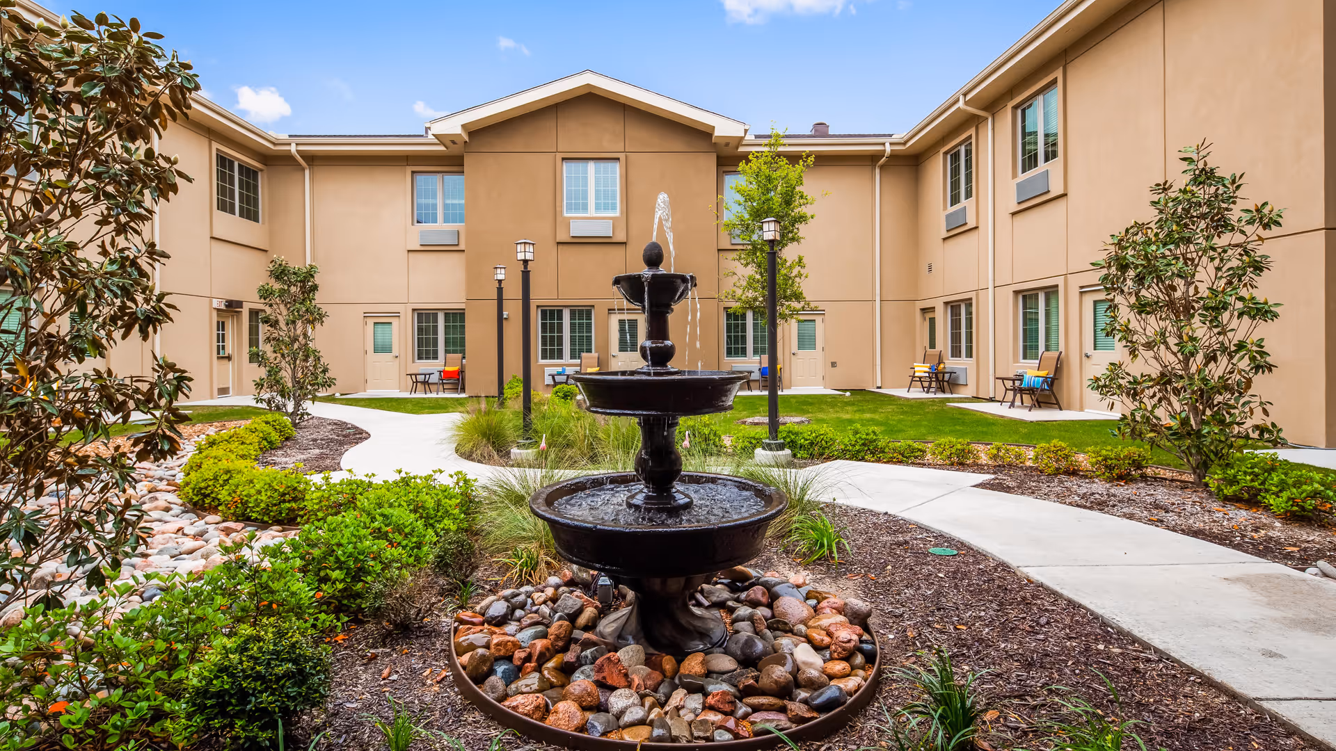 Outdoor courtyard area of a senior living facility with a three-tiered black water fountain surrounded by rocks and greenery. The courtyard is enclosed by a two-story beige building with multiple windows and doors. There are paved walkways, small trees, and shrubs, with benches placed near the building walls.