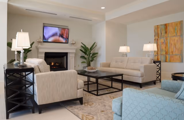 A cozy living room with a beige sofa and armchair, a blue patterned armchair, a black coffee table, and side tables with lamps. A fireplace with a TV mounted above it is in the background, flanked by two tall green plants. A large abstract painting hangs on the wall to the right.