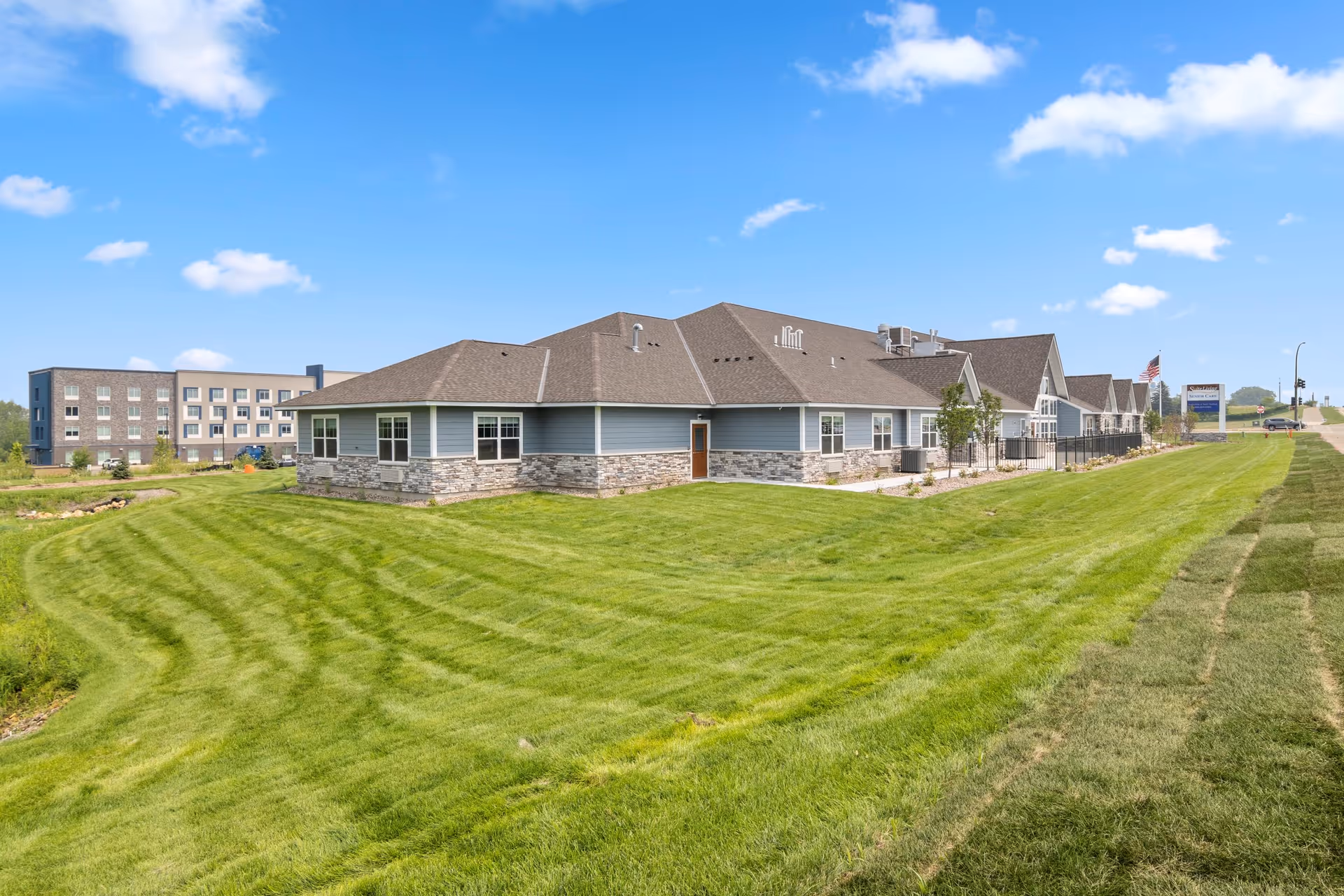 Exterior view of a senior living facility with a large, well-maintained green lawn in the foreground and a clear blue sky with a few clouds. The building has a combination of stone and blue siding with multiple windows and a brown roof. A sign and an American flag are visible near the road on the right side.