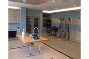 Bright multipurpose therapy/activity room with exercise equipment, a central table holding cleaning supplies, blue-striped walls and recessed ceiling lights.
