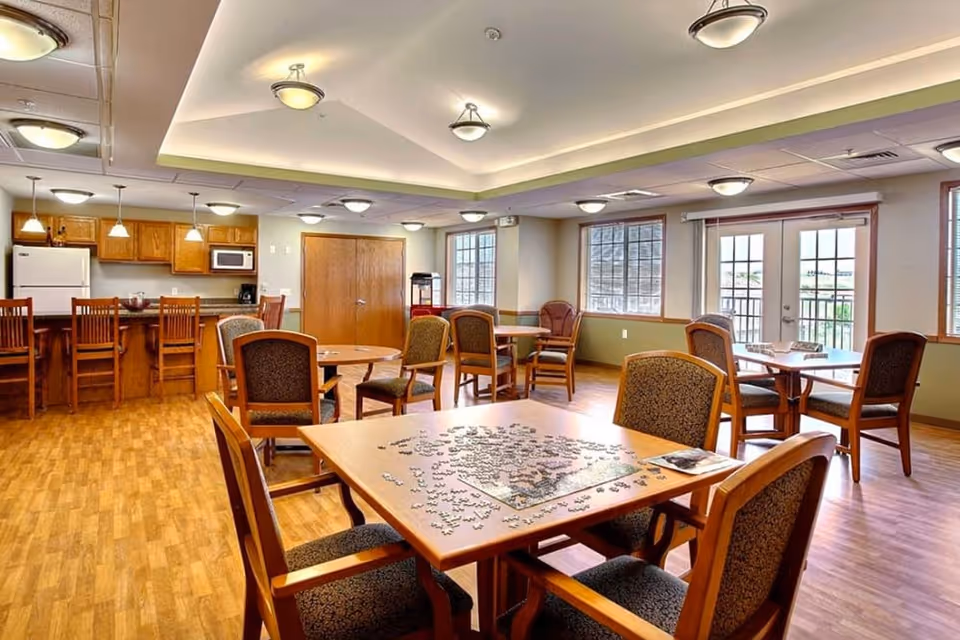A spacious common area in a senior living facility featuring multiple wooden tables and chairs. One table in the foreground has an incomplete jigsaw puzzle on it. The room has large windows and glass doors allowing natural light, wood flooring, and a kitchen area with wooden cabinets, a refrigerator, microwave, and bar stools in the background.