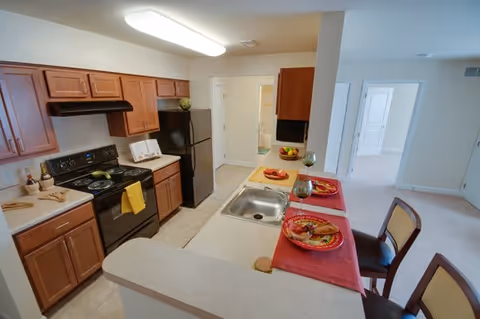 A clean and well-lit kitchen with wooden cabinets, a black stove and refrigerator, and a countertop with a sink. The countertop has two place settings with red placemats, plates of food, and glasses. The kitchen opens into a hallway and adjacent rooms with light-colored walls and carpeted floors.
