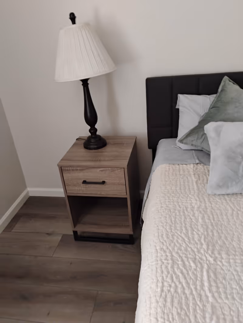 A bedroom corner featuring a wooden nightstand with a drawer and an open shelf, topped with a black lamp with a white pleated lampshade. Next to the nightstand is a bed with a dark upholstered headboard, light gray sheets, and multiple pillows in shades of gray and green. The floor is wooden and the walls are plain white.