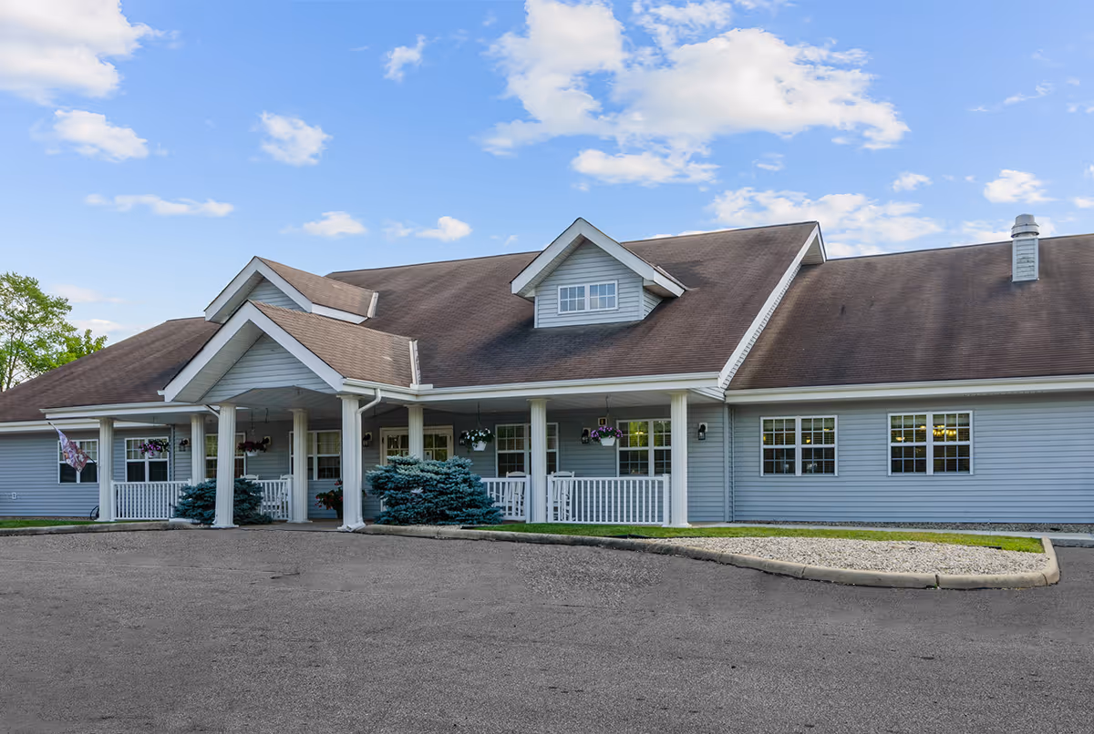 Front exterior of a single-story light-blue building with a covered porch, white columns, and a brown roof under a partly cloudy sky.