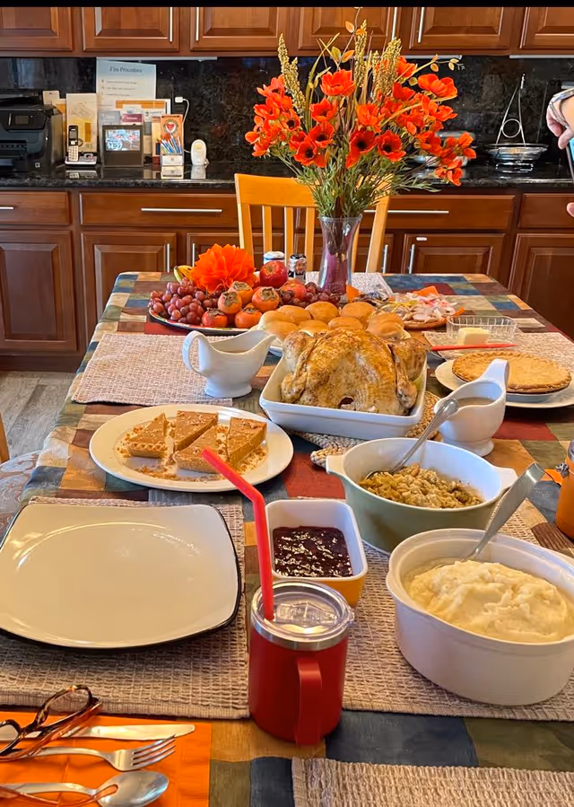 A dining table in a kitchen area laid out with a roasted turkey, side dishes, pies, rolls, fruit and a vase of orange flowers.