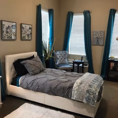 A neatly made bed with gray bedding and a patterned throw blanket in a bedroom. The room has three windows with teal curtains, a gray upholstered chair, a small side table with a laptop, and framed artwork on the walls.
