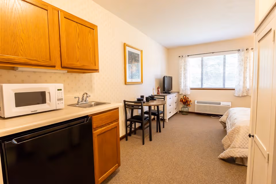 A small studio apartment with a kitchenette on the left featuring wooden cabinets, a microwave, a sink, and a mini refrigerator. In the middle, there is a small round dining table with two chairs. At the far end, a window with white curtains lets in natural light. Below the window is an air conditioning unit. To the right, there is a bed with a beige quilt and a white wardrobe. A TV sits on a white dresser next to a vase with flowers.
