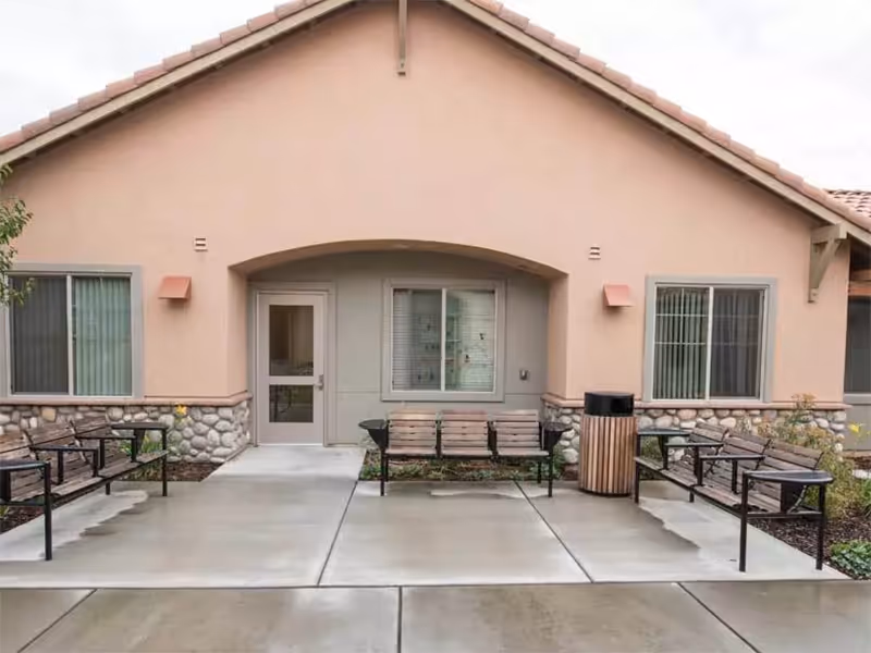 Exterior view of a single-story building with a beige stucco facade and stone accents at the base. There are two windows with vertical blinds and a door in the center under an arched entryway. In front of the building, there are three wooden benches with black metal frames arranged around a trash can on a concrete patio.