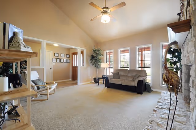 A spacious living room with beige carpet and walls, featuring a ceiling fan with light, a beige sofa, a tall plant next to a floor lamp, and large windows letting in natural light. There is a stone fireplace on the right with a decorative bird sculpture, and wooden shelves with various decorative items on the left. The room opens into a hallway with framed pictures on the wall and a wooden front door.