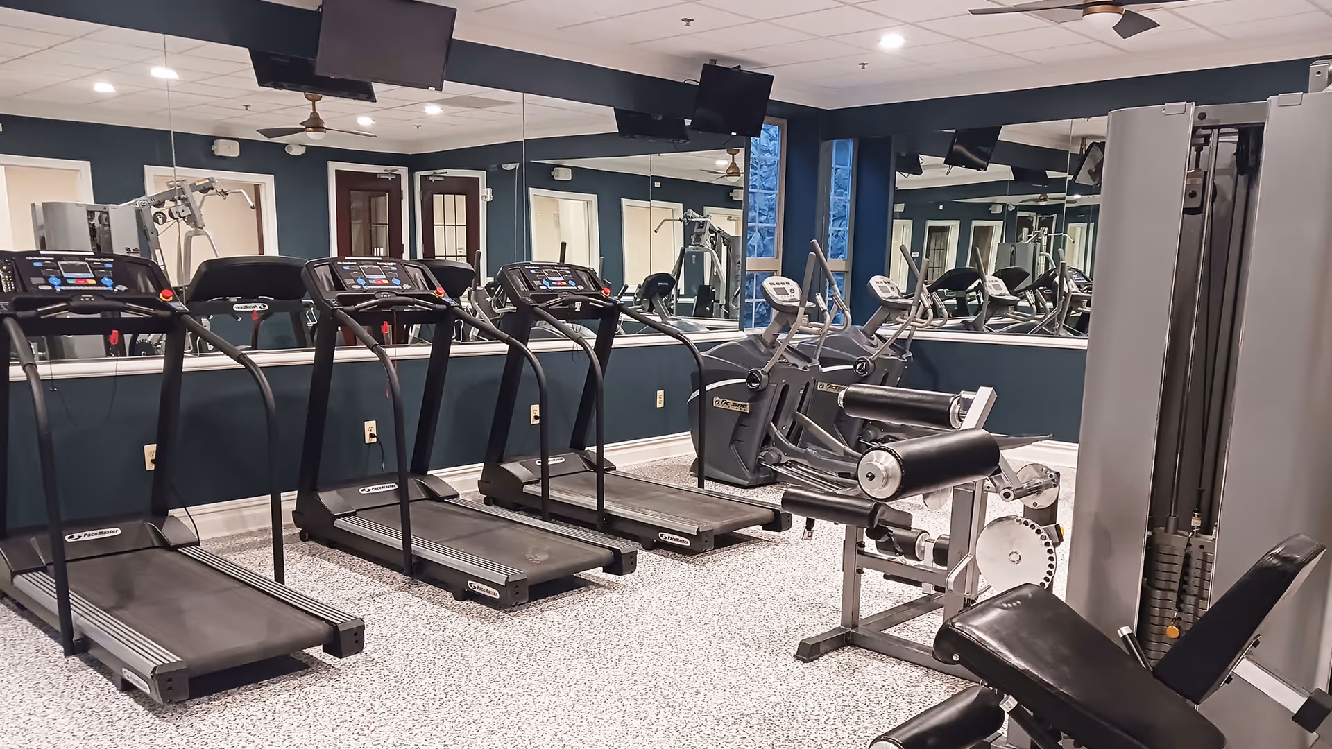 Indoor fitness room with multiple treadmills, exercise bikes, and weight machines. The room has large mirrors on the walls, a ceiling fan, and a speckled floor.
