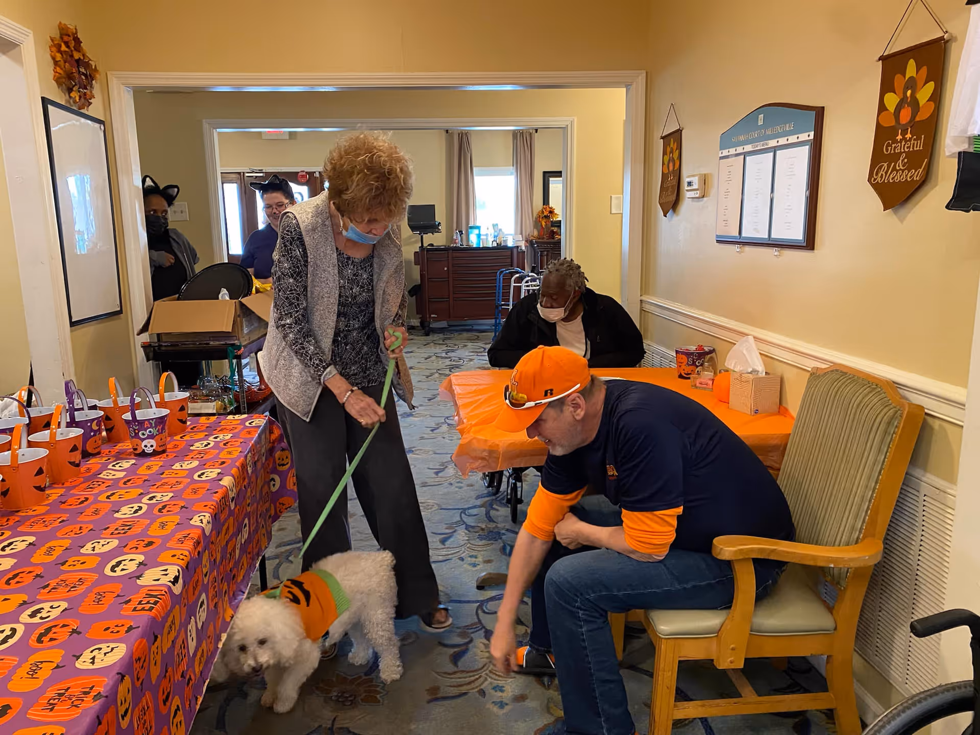 An indoor scene at Savannah Court of Milledgeville showing a woman holding a small white dog on a leash, a man sitting on a chair reaching towards the dog, and another person sitting at a table covered with an orange tablecloth. The room is decorated with Halloween-themed items including a table with a purple and orange Halloween tablecloth and small buckets, and wall hangings with autumn and Thanksgiving themes.