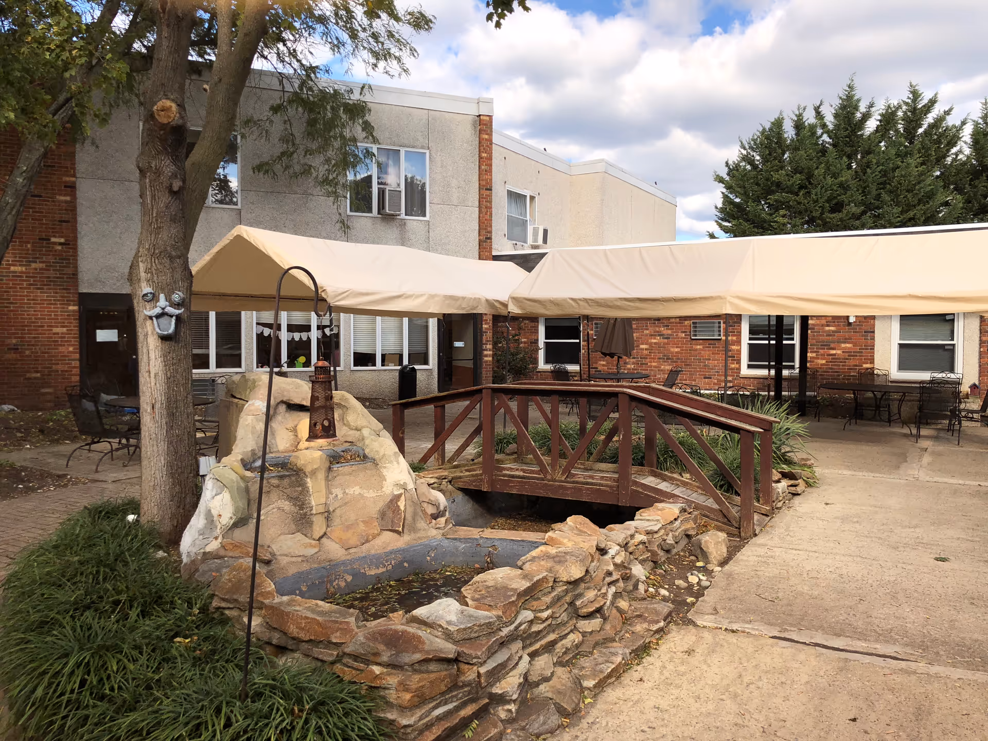 Outdoor courtyard area of a senior living facility with a small wooden bridge over a dry stone-lined pond, a tree with a decorative face attached, beige canopy coverings, and patio tables and chairs near a brick and concrete building under a partly cloudy sky.