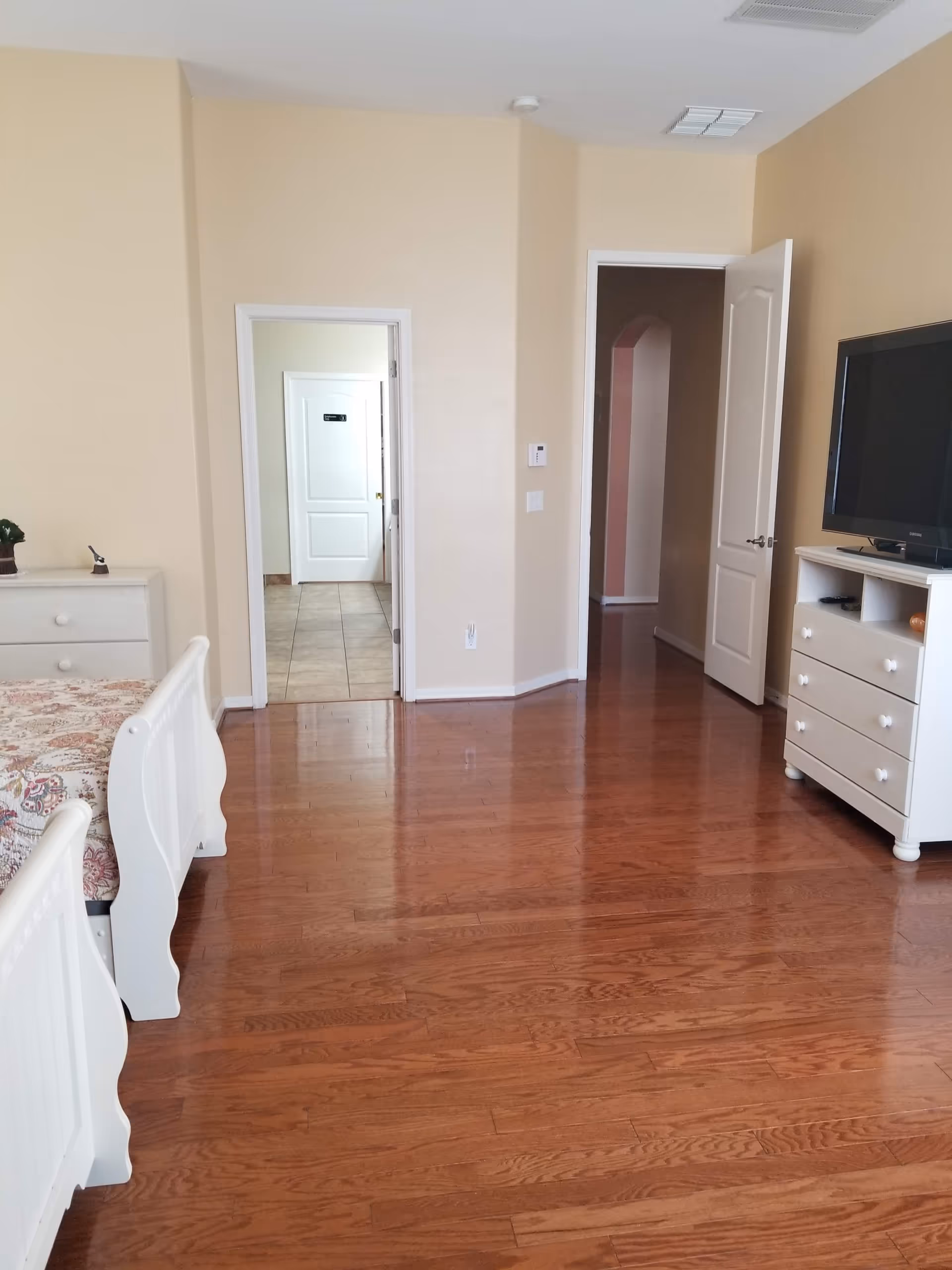 A bedroom with wooden flooring, a white bed with floral bedding on the left, a white dresser with a small plant on top, and a white TV stand with a flat-screen TV on the right. Two doorways are visible, one leading to a tiled area and the other to a hallway with an archway.