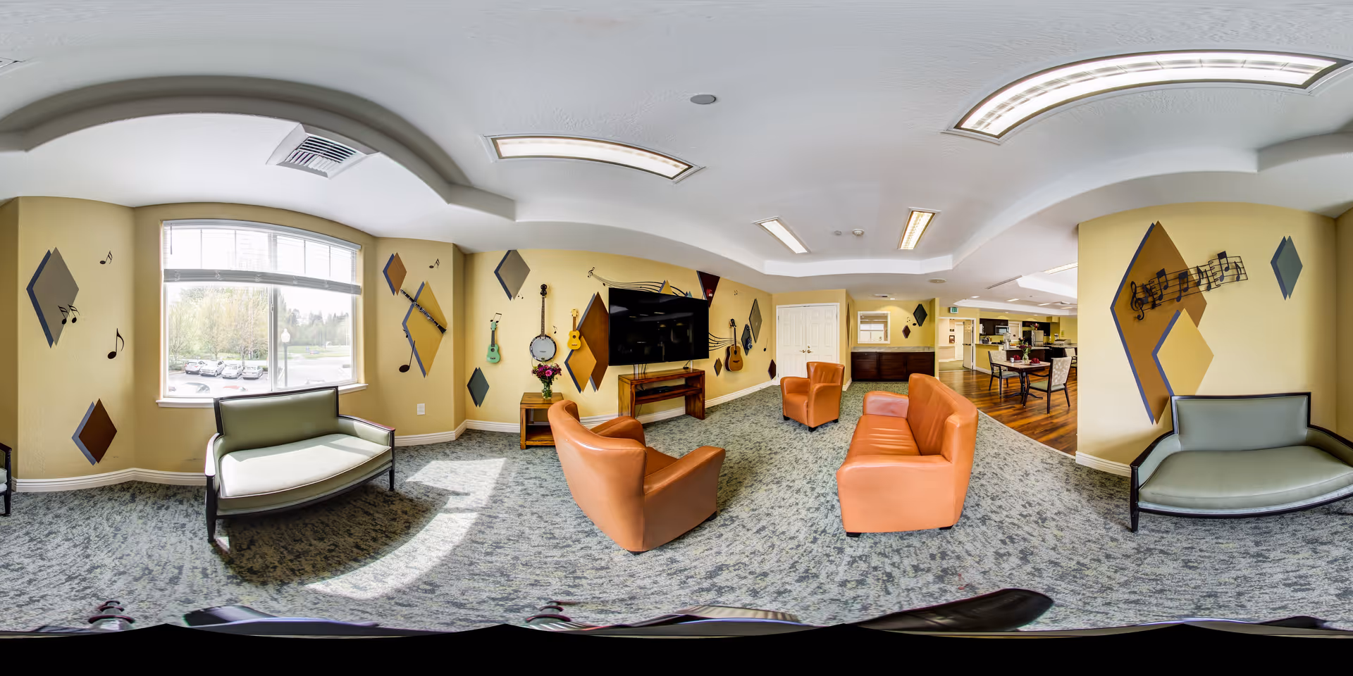 A spacious and well-lit living room area in a senior living facility with beige walls decorated with musical notes and instruments. The room features two orange armchairs, an orange sofa, and two green cushioned benches near the windows. A large flat-screen TV is mounted on the wall above a wooden stand, and the floor is carpeted with a patterned design. The room opens into a dining area with wooden flooring and tables with chairs.