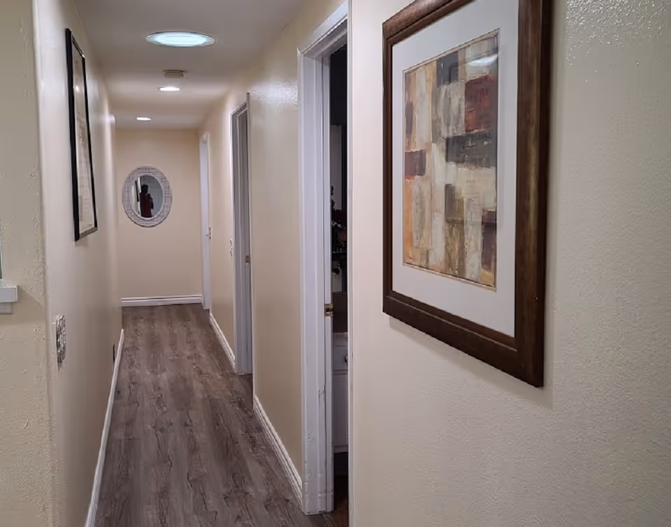 Narrow interior hallway with wood-look flooring, cream walls, recessed ceiling lights, framed artwork on the right and an oval mirror at the far end.