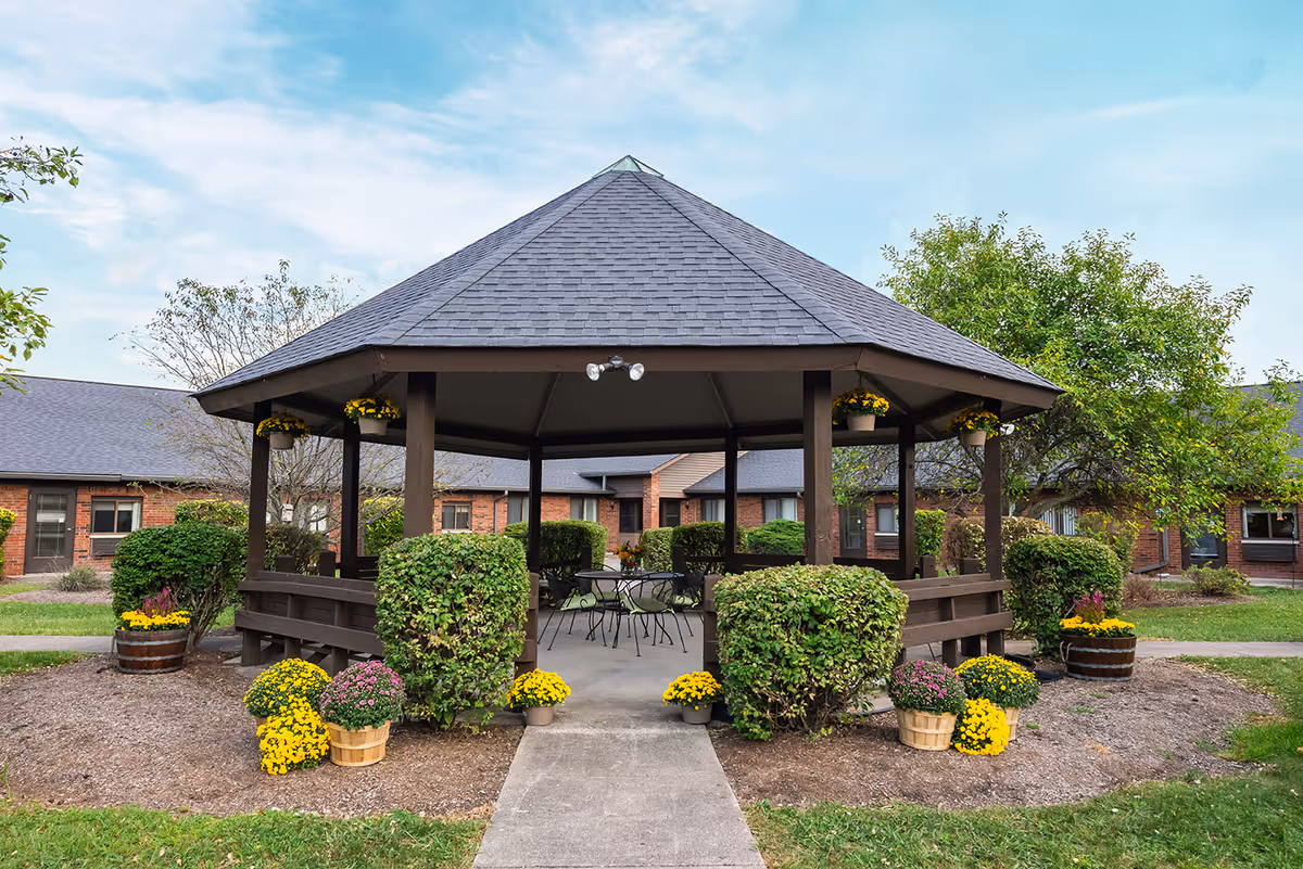A wooden gazebo with a dark shingled roof surrounded by neatly trimmed bushes and colorful potted flowers. The gazebo has metal chairs and tables inside and is situated in a garden area with a pathway leading to it. In the background, there are brick buildings and trees under a partly cloudy sky.