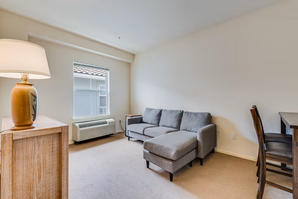 A small living room area with a gray sectional sofa, a wooden side table with a lamp, a window with a view of a tiled roof, and a wooden dining table with two chairs against a beige wall.