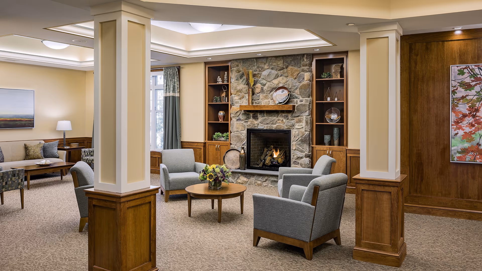 A cozy living room area in a senior living facility featuring a stone fireplace with a fire burning, surrounded by built-in wooden shelves with decorative items. Four gray armchairs are arranged around a round wooden coffee table with a flower arrangement. The room has beige walls, carpeted floor, wooden paneling, and two large pillars in the foreground.