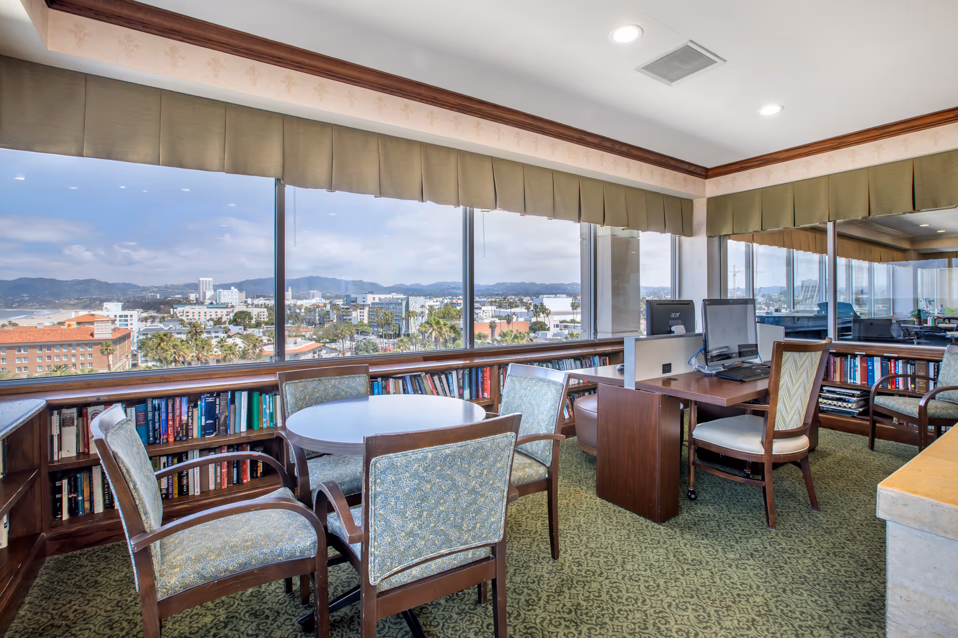 A bright room with large windows offering a panoramic view of a cityscape and mountains in the distance. The room features a round table surrounded by four upholstered chairs, a wooden desk with a computer, and bookshelves filled with books lining the walls beneath the windows. The carpet is green with a subtle pattern, and the ceiling has recessed lighting.