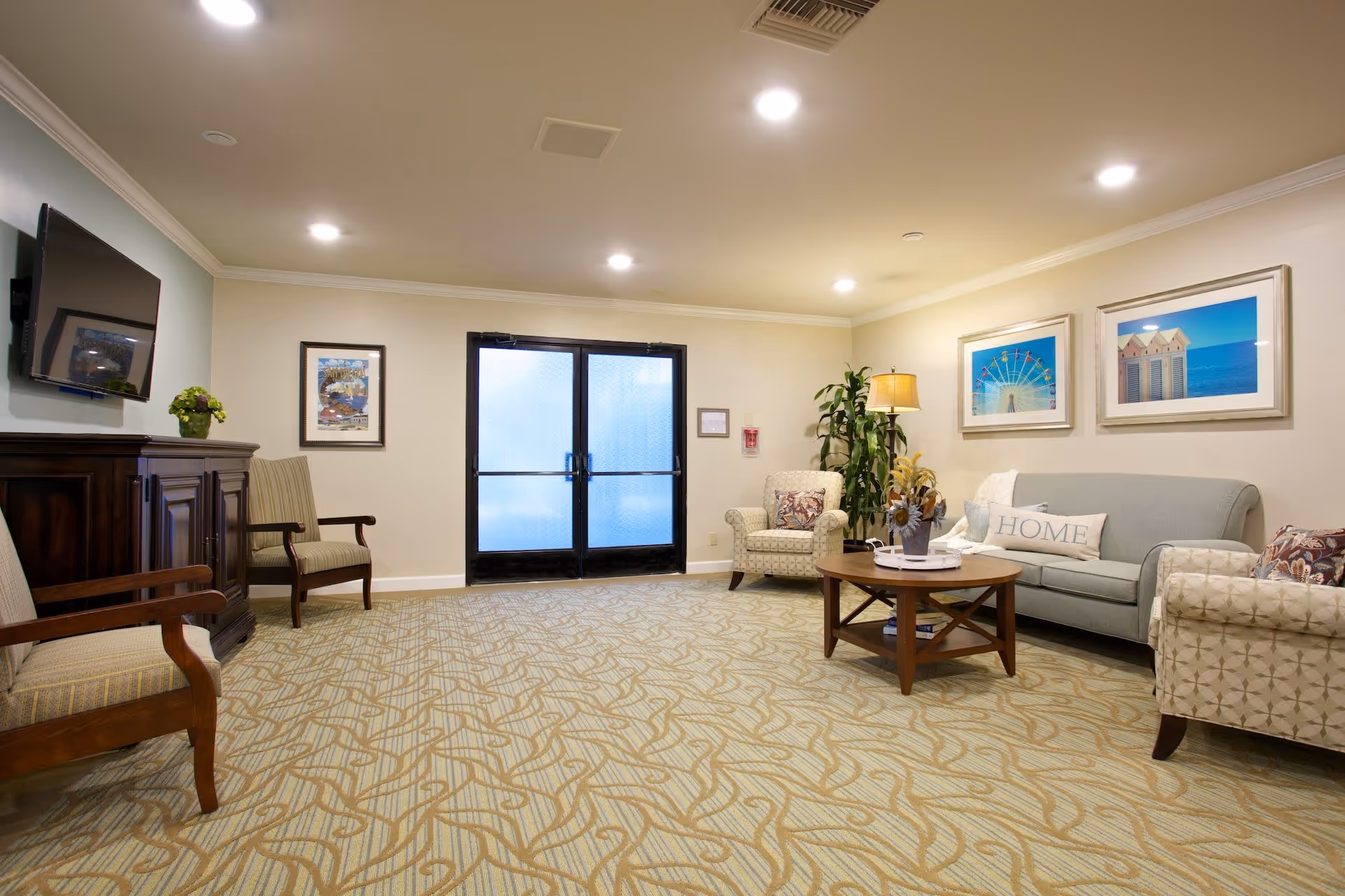 A cozy living room area with a patterned carpet, a light blue sofa with a 'HOME' pillow, two patterned armchairs, a wooden coffee table with a flower arrangement, a floor lamp, and framed pictures on the wall. There is a wooden cabinet with a flat-screen TV mounted above it and two wooden chairs with cushions. Double glass doors are visible in the background.