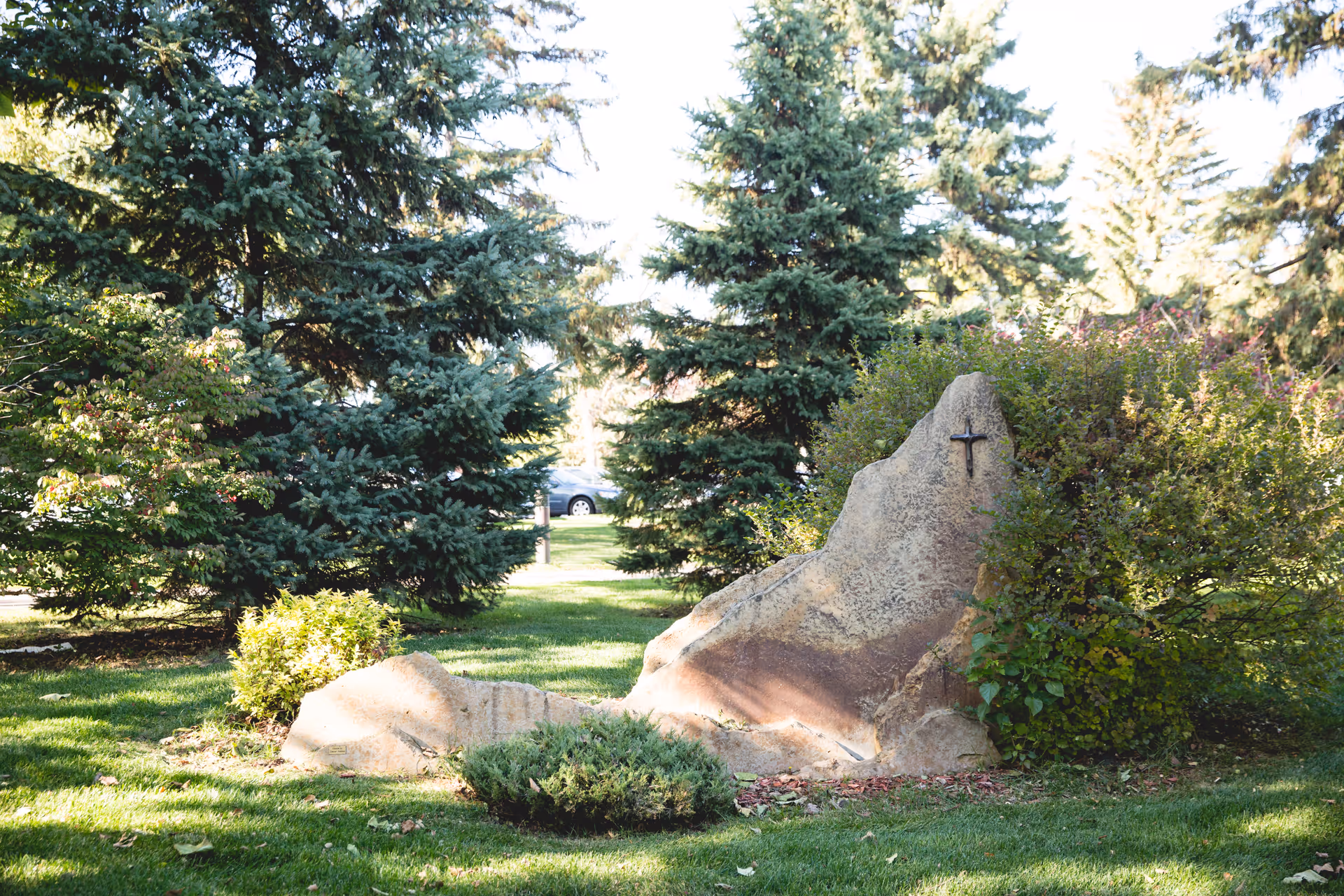 A landscaped outdoor area with green grass, bushes, and tall evergreen trees. There is a large rock with a small cross mounted on it, surrounded by greenery.