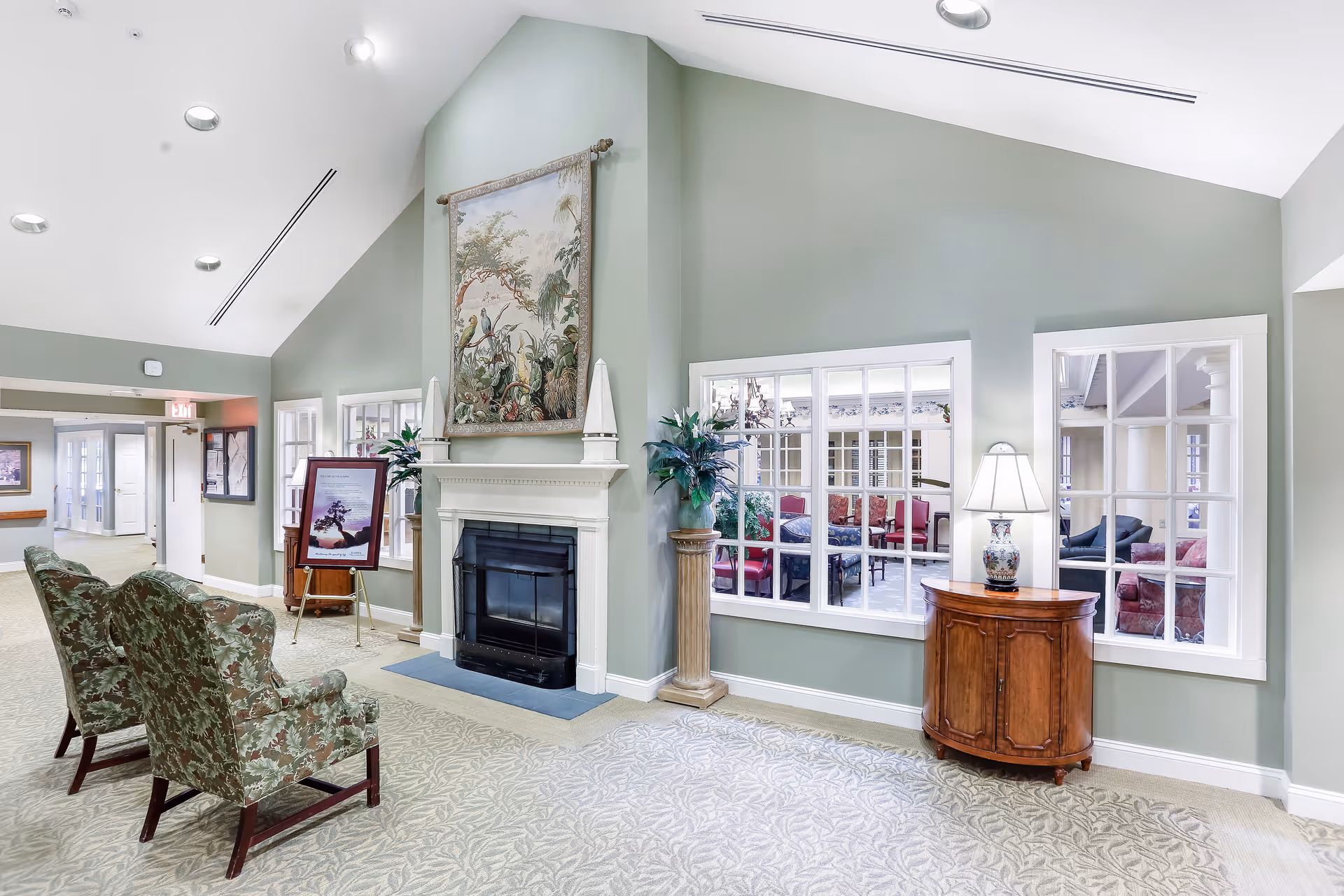 A bright and spacious interior hallway area with light green walls and a high vaulted ceiling. There is a white fireplace with a decorative tapestry above it, two floral upholstered chairs facing the fireplace, and a wooden cabinet with a lamp on it. Large windows on the right side reveal a cozy seating area with red and dark blue chairs. The floor is carpeted with a subtle leaf pattern.
