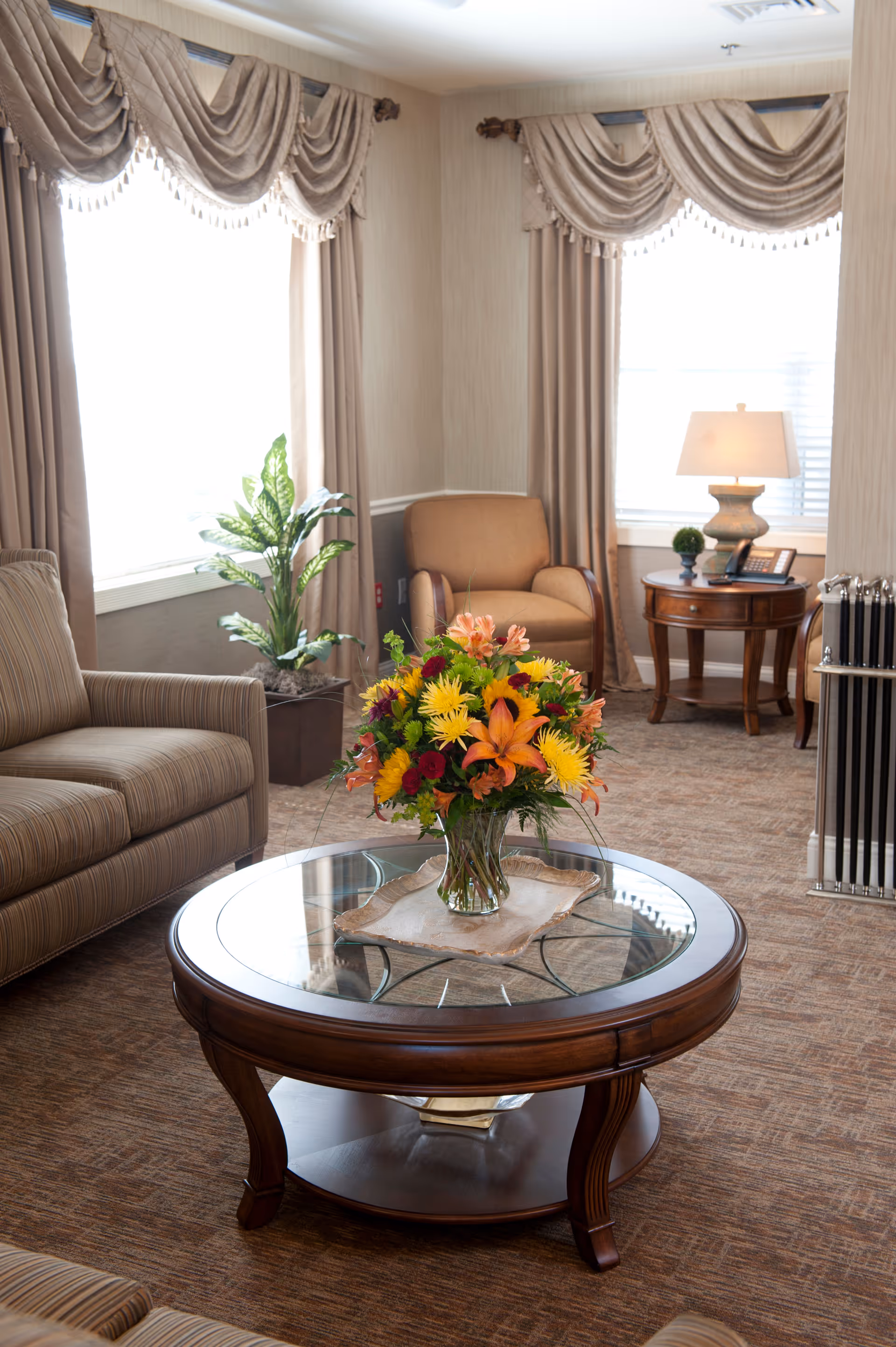 A cozy living room with a round glass-top coffee table holding a colorful floral arrangement, surrounded by upholstered chairs, a side table with a lamp, and draped windows.
