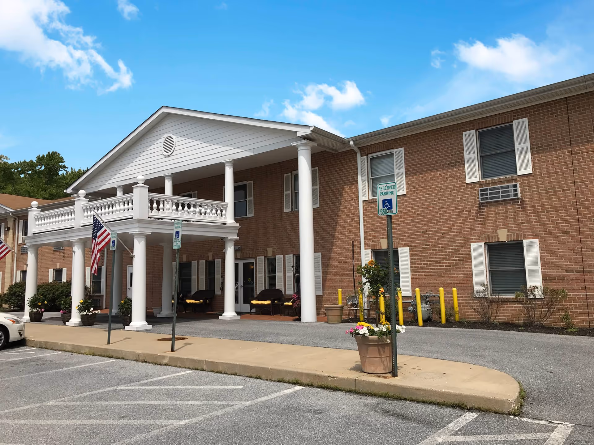 Brick two-story assisted living building with a white columned entrance, American flags, and parking spaces including handicapped signs.