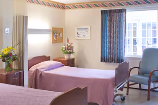 A bright shared patient bedroom with two beds, bedside tables with flowers, a chair, and a window.