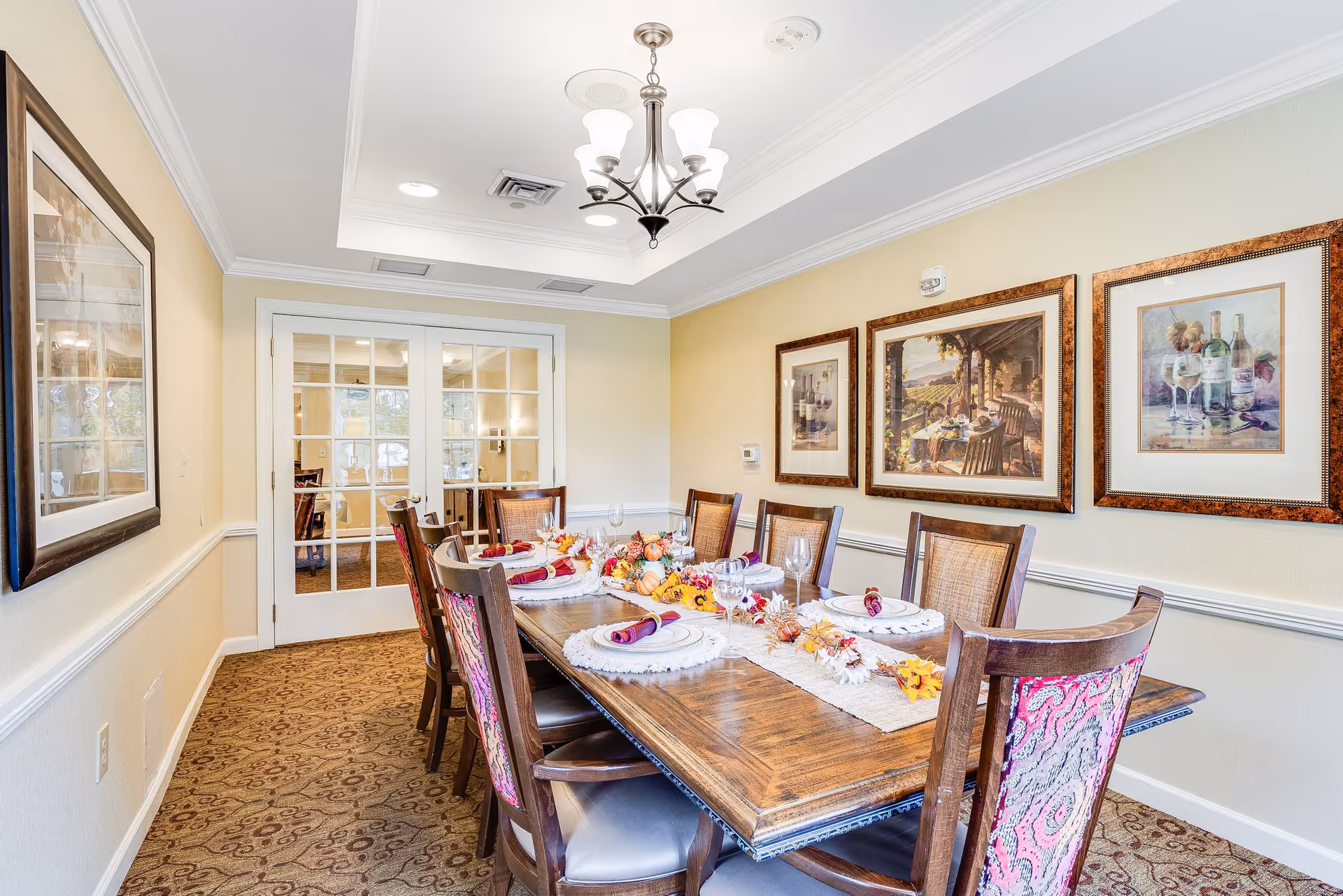 A well-lit dining room with a long wooden table set for eight people. The table is decorated with a fall-themed centerpiece and place settings including plates, wine glasses, and napkins. The chairs have wooden frames with patterned fabric backs. The room has beige walls adorned with framed artwork depicting wine and vineyard scenes. French doors with glass panes lead to another room.