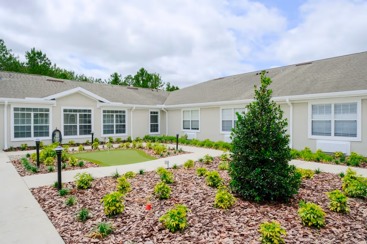 Outdoor courtyard area of a senior living facility with a small putting green, landscaped garden beds with mulch and shrubs, surrounded by a single-story building with multiple windows and a gray roof under a cloudy sky.