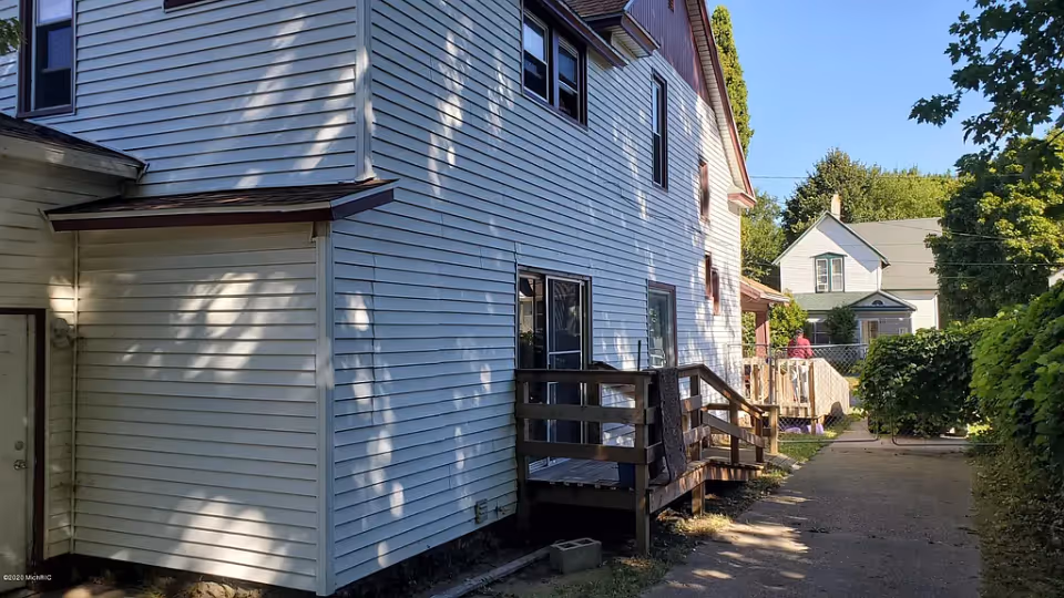Side exterior of a white vinyl-sided two-story house with a small wooden deck and a driveway leading to neighboring homes.