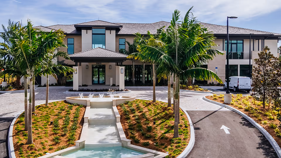 Front exterior view of the Shell Point Welcome Center building with a paved driveway, landscaped garden with palm trees, and a small water feature leading up to the entrance under a covered porch.