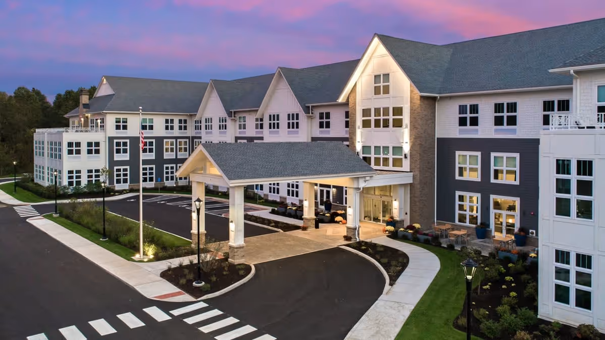 Exterior view of a large, modern senior living facility building at dusk with a covered entrance, well-lit pathways, landscaped greenery, and a parking area. The sky is a gradient of purple and pink hues.