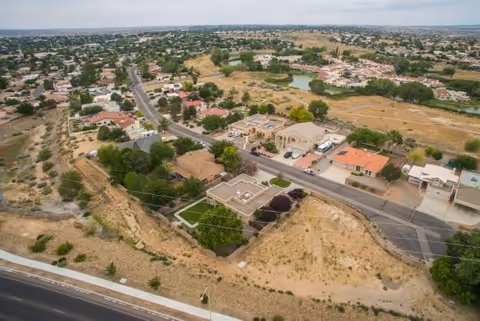 Aerial view of a suburban neighborhood with houses, roads, and some greenery. The area includes a mix of residential buildings and open land with sparse vegetation.