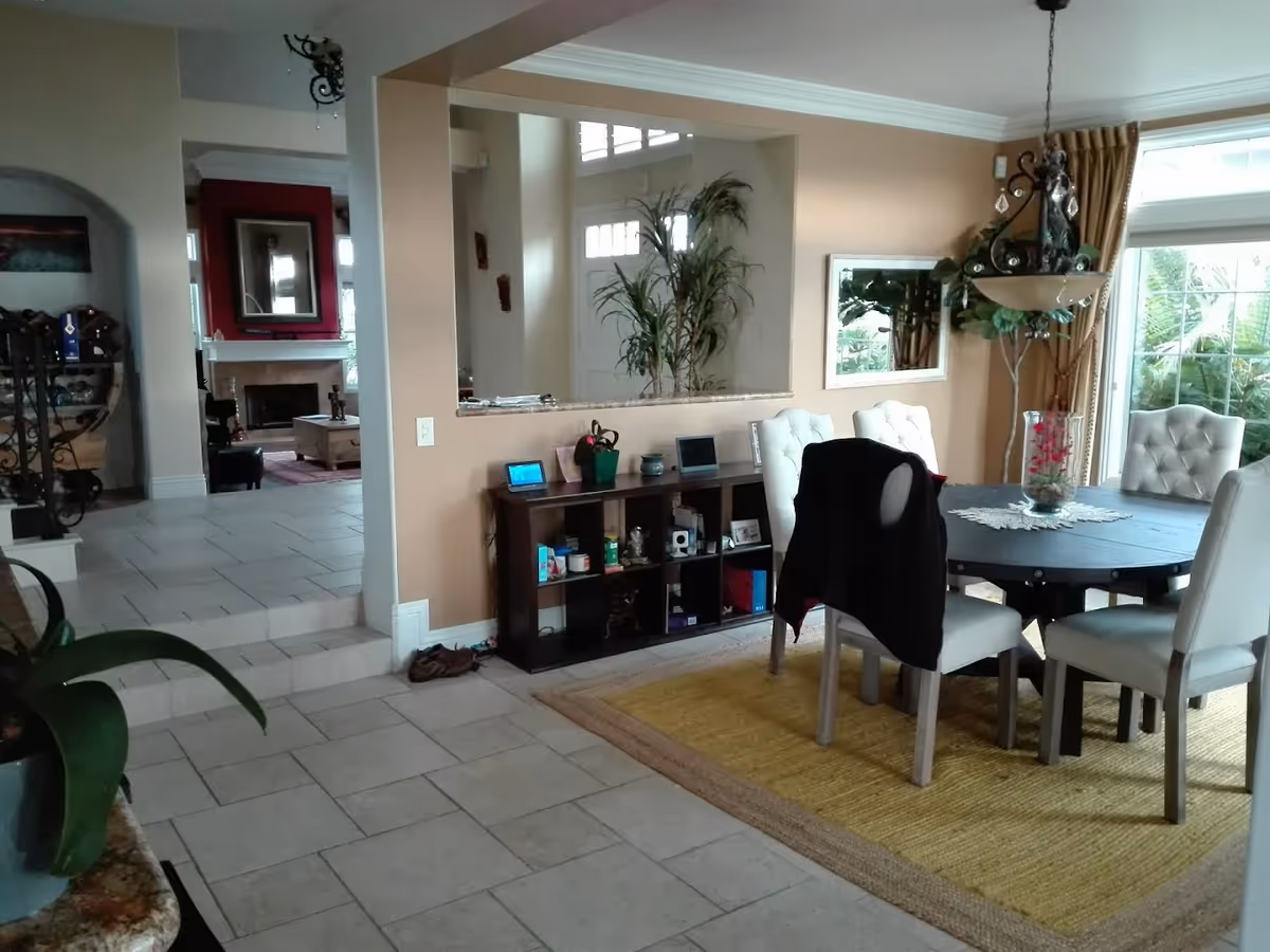 Interior view of a residential dining area with a round black dining table surrounded by six white cushioned chairs, one with a black jacket draped over it. The room has beige walls, a large window with curtains, a hanging light fixture, and a yellow area rug under the table. Adjacent to the dining area is a small shelving unit with various items and a large mirror above it. In the background, there is a living room area with a fireplace, a red accent wall, and a coffee table. The floor is tiled throughout.
