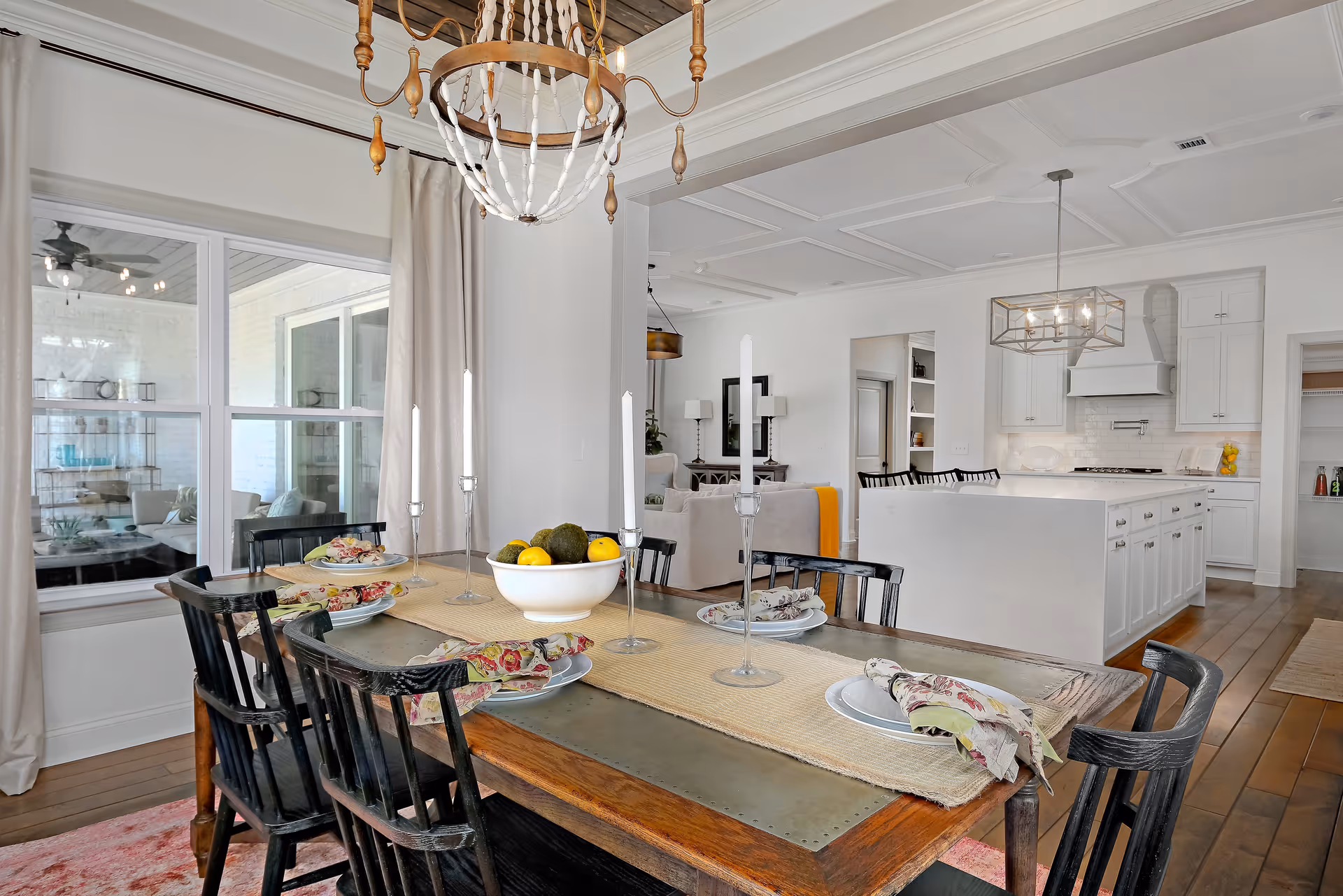 A bright and spacious dining area with a wooden table set for four, featuring floral napkins and tall white candles. The room has large windows with beige curtains, and a chandelier hanging above the table. In the background, there is a modern white kitchen with an island, bar stools, and pendant lighting, as well as a glimpse of a living room area with sofas and lamps.