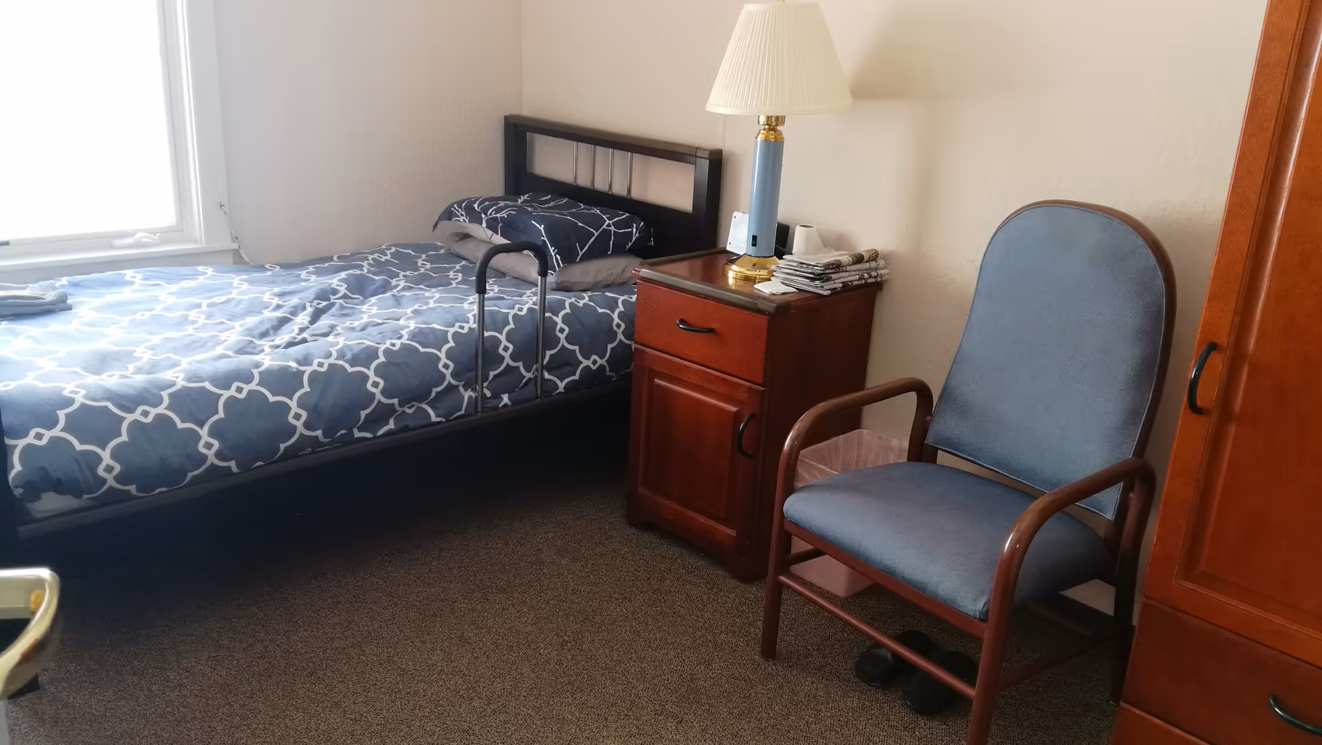 Simple single-occupancy bedroom with a twin bed, bedside table with a lamp, and an upholstered chair.