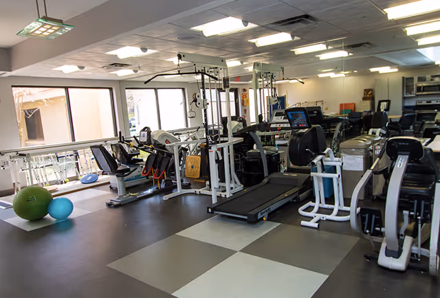A bright exercise/physical therapy room with treadmills, bikes, weight machines, and exercise balls.