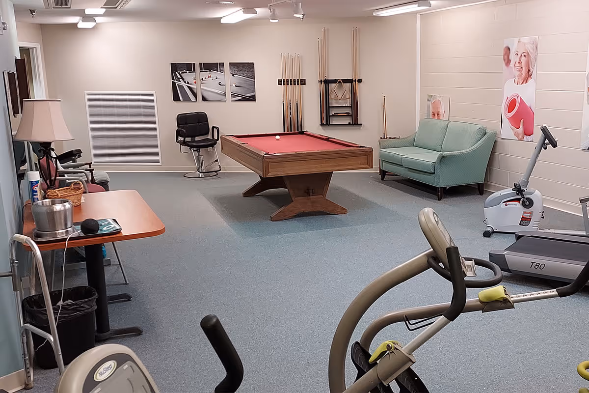 A recreational room featuring a pool table with a red felt top in the center, exercise equipment including a stationary bike and treadmill, a green couch against the wall, and a table with a lamp and various items on it. The walls have pictures of elderly people and pool cues are mounted on the wall behind the pool table.