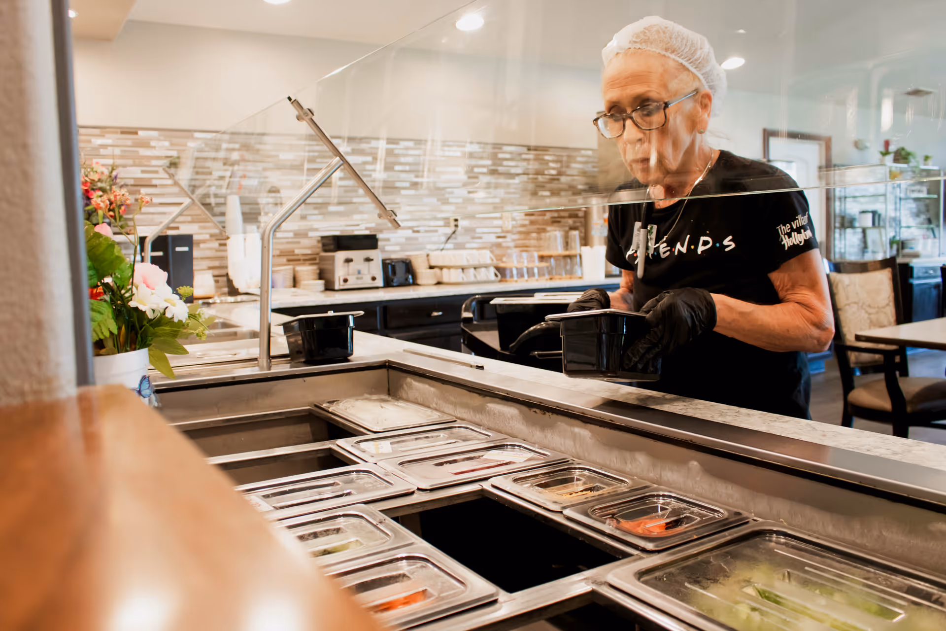 A woman wearing a hairnet, glasses, and black gloves is serving food from a buffet-style salad bar in a dining area. The background shows a kitchen counter with appliances and a tiled backsplash, along with tables and chairs for dining.