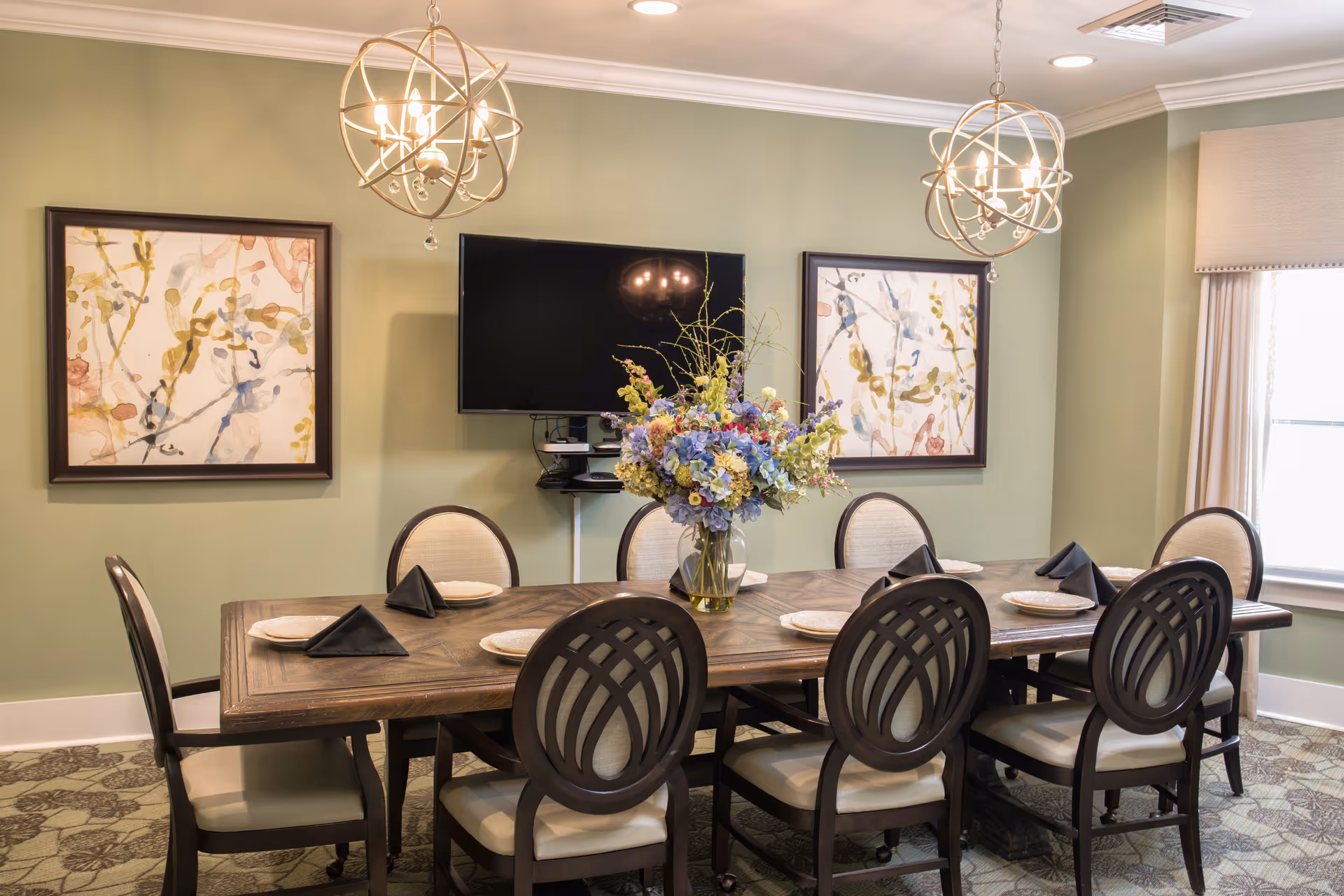 A dining room with a long wooden table set with white plates and black folded napkins. Eight cushioned chairs with dark wooden frames surround the table. A large floral centerpiece in a glass vase is placed in the middle of the table. Two modern spherical chandeliers hang from the ceiling. The walls are painted light green and decorated with two abstract floral paintings. A flat-screen TV is mounted on the wall between the paintings. A window with beige curtains allows natural light into the room.
