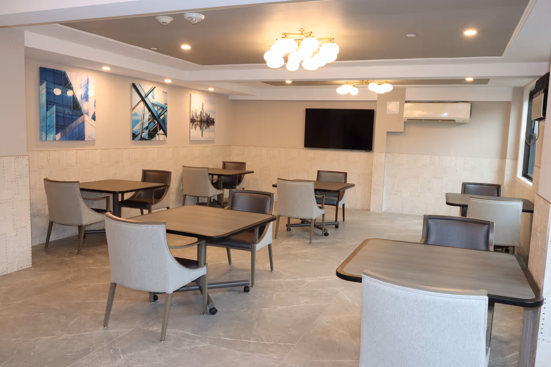 A bright communal dining room with multiple tables and upholstered chairs, wall art, a mounted TV, and ceiling lights.