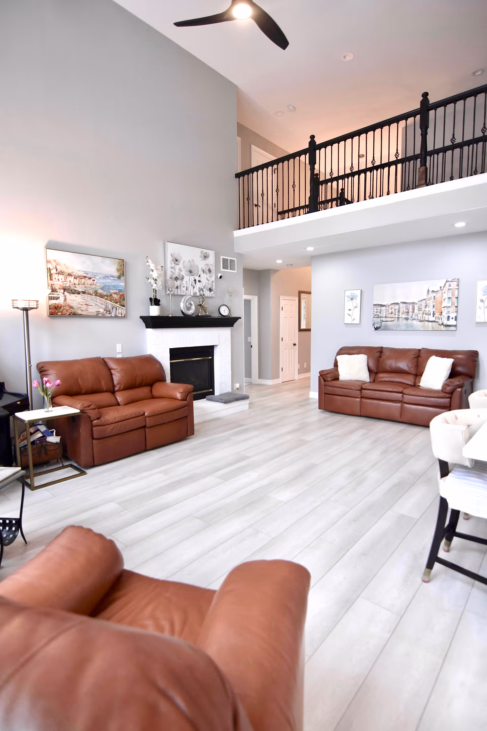 A spacious living room with high ceilings and light-colored wooden flooring. The room features three brown leather sofas, a white fireplace with decorative items on the mantel, and artwork on the walls. There is a black ceiling fan above and a balcony with black railing overlooking the room.