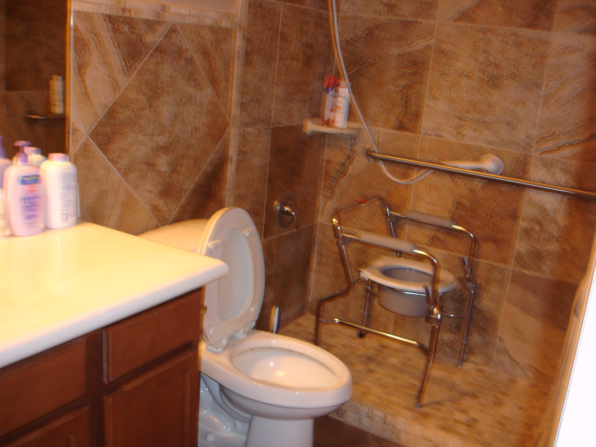 Bathroom with brown tiled walls and floor, a white toilet with the lid open, a vanity with a white countertop and several bottles of lotion or soap, a shower area with a handheld showerhead, a grab bar, and a metal shower chair with armrests.